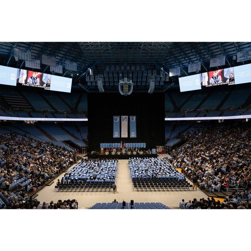 Long-range angle of the crowd at U.N.C. Chapel Hill's Winter Commencement at the Dean E. Smith Center. Seen are graduates sitting in rows on the floor, guests filling the lower bowl of the arena, and a stage at the front.